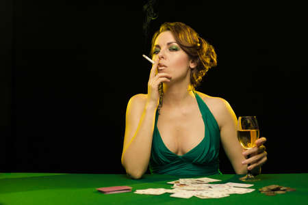 A young lady in a green dress smokes a cigarette and blows smoke and plays cards on a table on green clothの写真素材