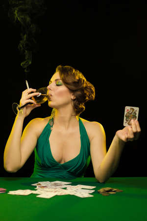 Woman in a green dress smokes a cigarette and blows smoke and plays cards on a table on green clothの写真素材