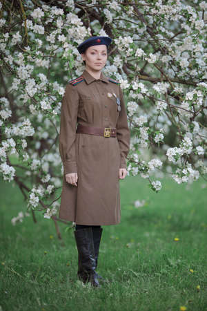 A young female pilot in uniform of Soviet Army pilots during the World War II. Military shirt with shoulder straps of a major and a beret. In spring blooming apple orchardの写真素材