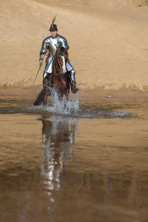A young adult man in knightly armor rides a horse on a river along a sandy shoreの写真素材