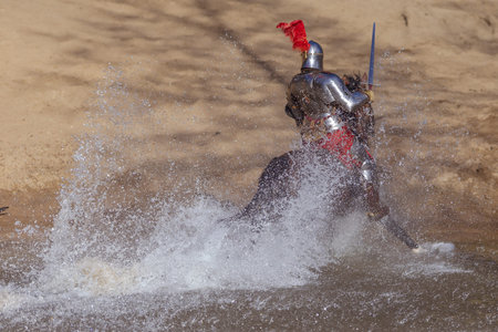 A young adult man in knightly armor rides a horse on a river along a sandy shore