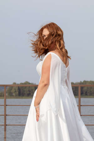 Red-haired bride in a white dress posing on a quay near a lakeの写真素材