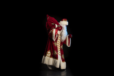Santa Claus in a red fur coat, with a white beard and a bag of gifts posing on a black background