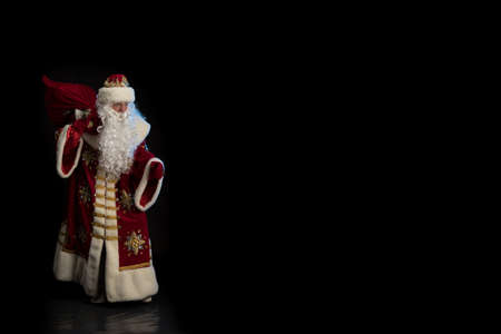 Santa Claus in a red fur coat, with a white beard and a bag of gifts posing on a black backgroundの写真素材
