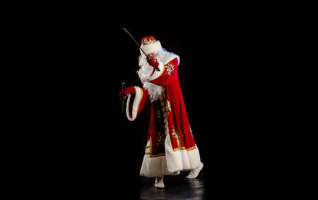 Santa Claus in a red fur coat, with a white beard with two swords in his hands, posing on a black backgroundの写真素材