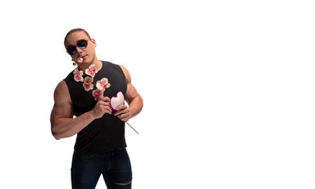 Portrait of a brutal man bodybuilder athlete in sunglasses with a sprig of blooming pink orchid and soft heart in the hands on a white backgroundの写真素材