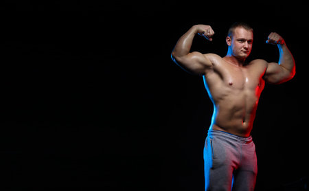 male bodybuilder athlete with naked torso posing against a black background, in red and blue light