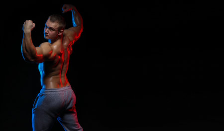 male bodybuilder athlete with naked torso posing against a black background, in red and blue light
