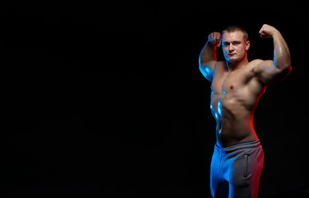 male bodybuilder athlete with naked torso posing against a black background, in red and blue light