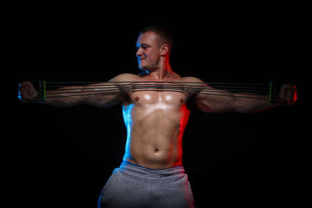 male bodybuilder athlete with naked torso doing exercises with expander on a black background, in red and blue lightの写真素材