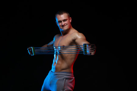 male bodybuilder athlete with torso doing exercises with expander on a black background, in red and...