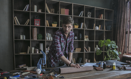 Young man leather maker unfolds roll of leather on table. Small business at homeの写真素材