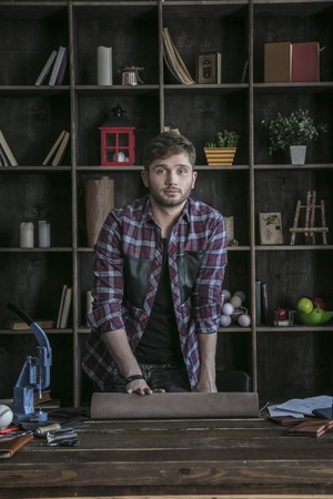Young man leather maker stand near wooden table with tools and roll of leatherの写真素材