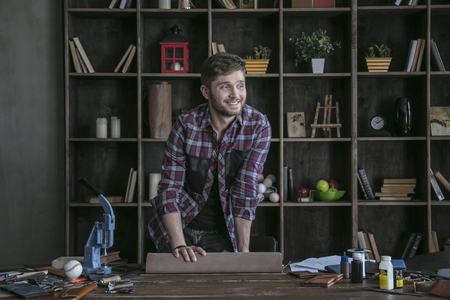 Young man leather maker hold roll of leather on wooden table with toolの写真素材