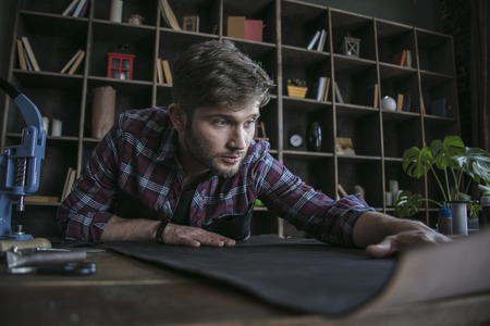 Young man sitting at wooden working table with piece of leather. Leather makerの写真素材