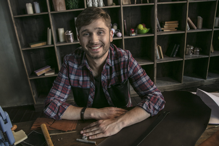 Young man leather manufacturer smiling and sitting at wooden table with toolsの写真素材