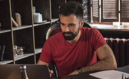 Young businessman with beard sitting in chair and working on laptop on tableの写真素材