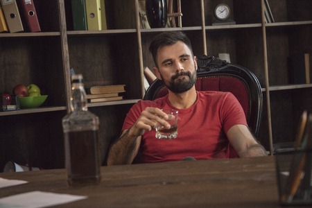 Young bearded businessman sitting in office chair with glass of boozeの写真素材