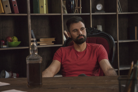 Young bearded businessman sitting at table with bottle of expensive alcoholの写真素材