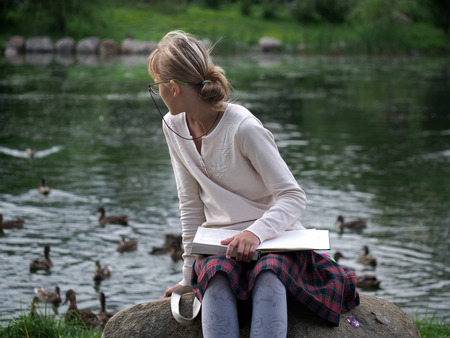 Portrait of a schoolgirl. Girl in glasses with a book in a city park. Pond, green trees. The girl looks at ducksの写真素材