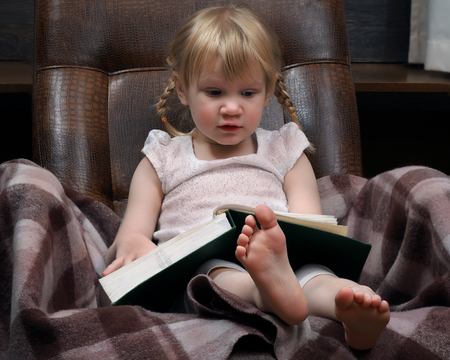 Little girl with a large book. Green Book. The girl in the chair. A girl with two braids, wearing a white dress, barefoot. The plaid chair. Girl learns to read. Portrait of a young childの写真素材