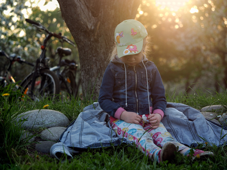 Cycling. A child sitting on the jacket resting in the grass. Girl considers flower dandelions. Nearby are the bikes. Evening, sunset, sun rays. Park in the cityの写真素材