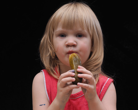 The child is eating a pickle. Portrait of the girl in a pink T-shirt with pickles. Black background. On the nutrition of children. small childの写真素材