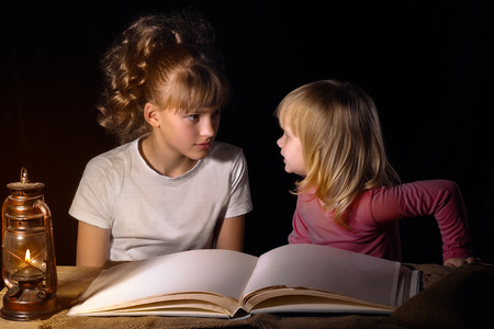 Two sisters tell each other scary stories at night. Open book, vintage lamp (kerosene). Black background. A family. Girls sister, older and one younger. Babies interesting and scary. Bedtime Storyの写真素材