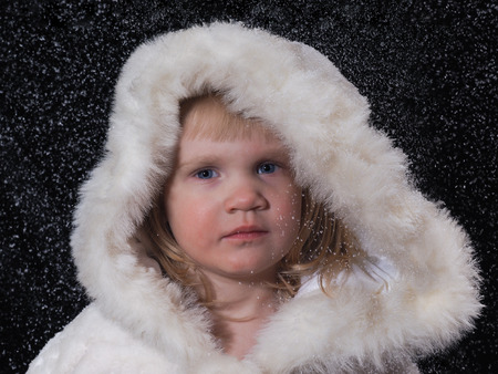 The little girl in a white fur coat under the falling snow. Face close, on the face of snow. Background black. Girl Snow Maidenの写真素材