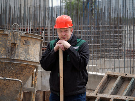 Tired working on a construction site. Home construction. A man in a helmet and overalls holding a shovel. Rest from workの写真素材