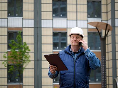 Engineer standing on the background of a new apartment building. House is built. Engineer in a helmet with papers in hand on the phone. The concept of new housing construction, real estateの写真素材