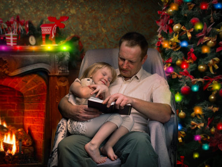 Christmas story by the fireplace. Dad reads a book a little daughter. Christmas tree, festive interior of the house. Christmas Eveの写真素材