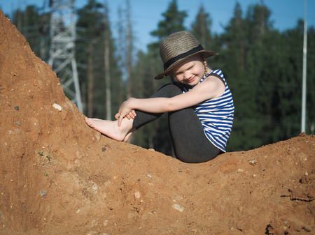 The child is sitting on a mountain of sand. Blue sky. A little girl, wearing a hat and barefoot. Journey with the concept of childrenの写真素材
