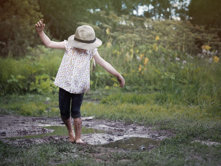 A child playing in a muddy puddle. Dirty girl in a hat and barefoot. Rural road