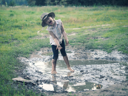 A child playing in a muddy puddle. Dirty girl in a hat and barefoot. Rural road