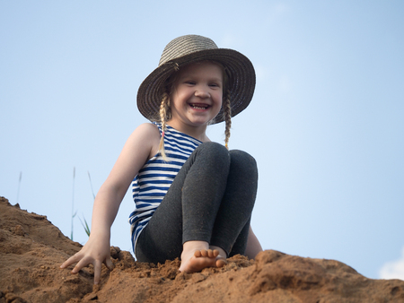 The child is sitting on a mountain of sand. Blue sky. A little girl, wearing a hat and barefoot. Journey with the concept of childrenの写真素材