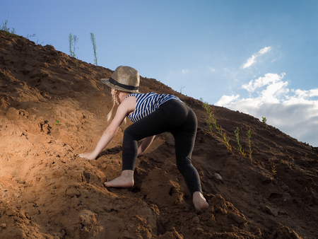 Child is climbing a mountain of sand. A little girl, wearing a hat and barefoot. Journey with the concept of childrenの写真素材