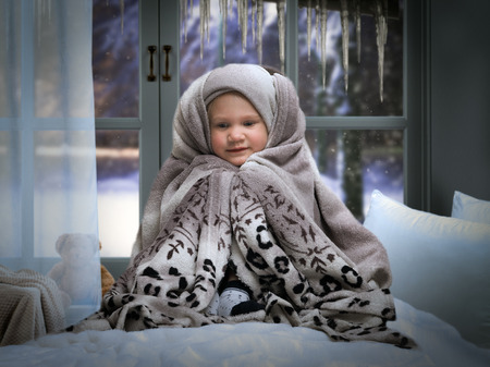 small child wrapped in a blanket on the bed. Children's room window. Outside the window, snow, winterの写真素材