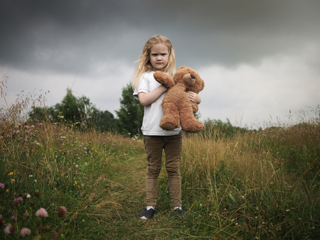 small child is on the field with a Teddy bear. Ominous sky. The concept of children's loneliness, stress, depressionの写真素材