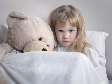 Sad little girl with plush bear in bed. Portrait of a distressed child. The concept of children's loneliness, illnessの写真素材