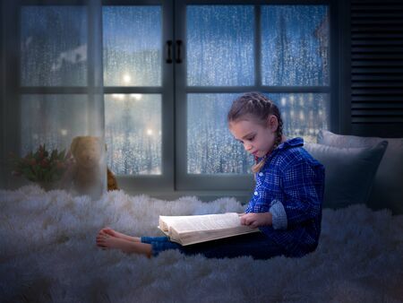 Little girl with a book late at night in a cozy children's room on the bedの写真素材