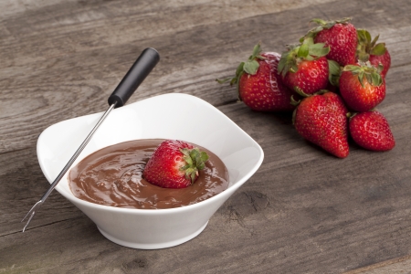 Vertical image of a bowl of chocolate dip and fondue stick beside a pile of strawberries laid in a wooden tableの写真素材