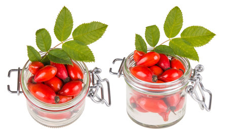 Two jars with red rosehips and green leaves. Fresh ripe hips and brier twig in glass. Isolated on...