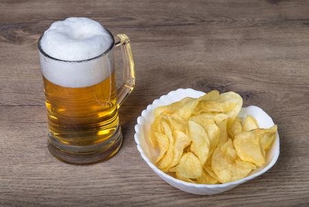 Beer pint and crispy potato chips on a wood table. Light alcoholic lager in the glass mug and pile of fat fried crisps in a white bowl.の写真素材