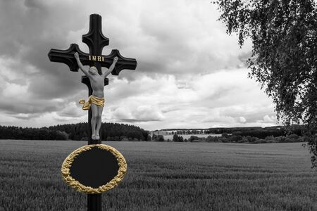 Jesus Christ crucifix in black-white rural landscape with a cornfield and dramatic sky. Christian faith. Vintage metal religious cross closeup. Copy space in oval gold frame. Dark mourning background.の写真素材