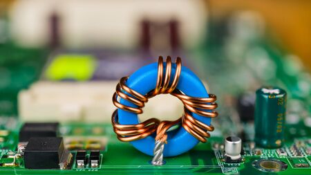 Blue toroidal induction coil on green circuit board. Computer mainboard detail. Electronic components. Ferrite core inductor, capacitor, transistor or diode. Blurry background, bokeh. Selective focus.の写真素材