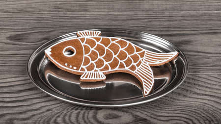 Sweet fish shaped gingerbread cookie with reflection on round silver plate. Closeup of Xmas pastry decorated with white icing on shiny stainless serving salver. Cute baked child toy on chrome platter.の写真素材