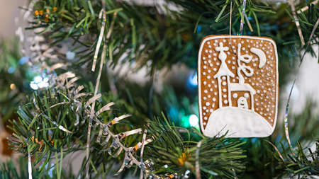 Sweet gingerbred and silvery string on needles of ornate artificial Christmas tree. Closeup of traditional baked Xmas cookie with painted snowy Christian church from white icing hanging on green twig.の写真素材