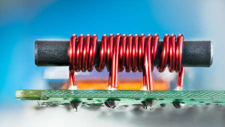 Electronic transformer with ferromagnetic core and coil copper wire on PCB. Closeup of red cylindric inductor soldered on green circuit board with tin on bottom side and blue background. Cable TV amp.の写真素材