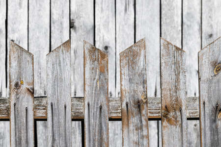 Wood fence from pointed boards with pale wooden building wall in blur background. Closeup of weathered safety fencing with gaps between brown planks with faded white paint coating.の写真素材